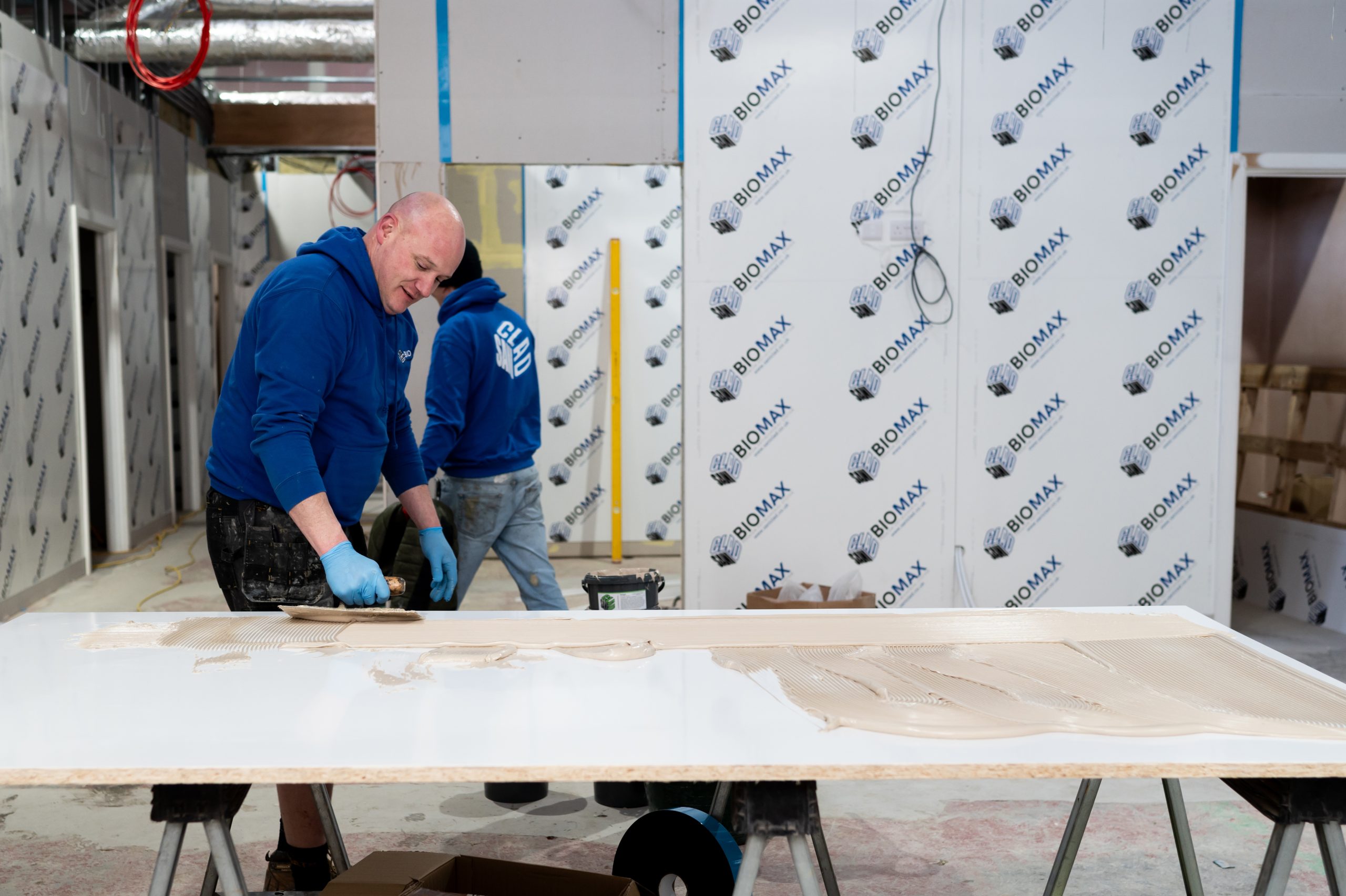 Two workers in blue hoodies working in a construction area covered in SANICLAD BIOMAX cladding. One worker is applying adhesive to a large panel on a workbench, while the other is standing in the background.